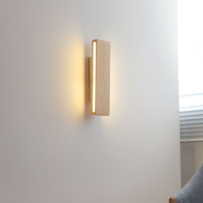 Vertical wooden wall lamp glowing on a white wall beside a window with blinds and part of a grey armchair. 15