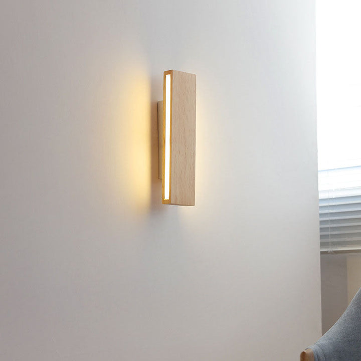 Vertical wooden wall lamp glowing on a white wall beside a window with blinds and part of a grey armchair. 15