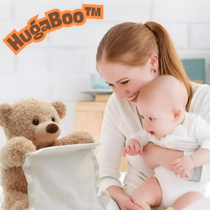 Uśmiechnięta kobieta z niemowlęciem na rękach patrzy na beżowego pluszowego misia, który wygląda zza białej chusteczki w zabawie w chowanego. 1