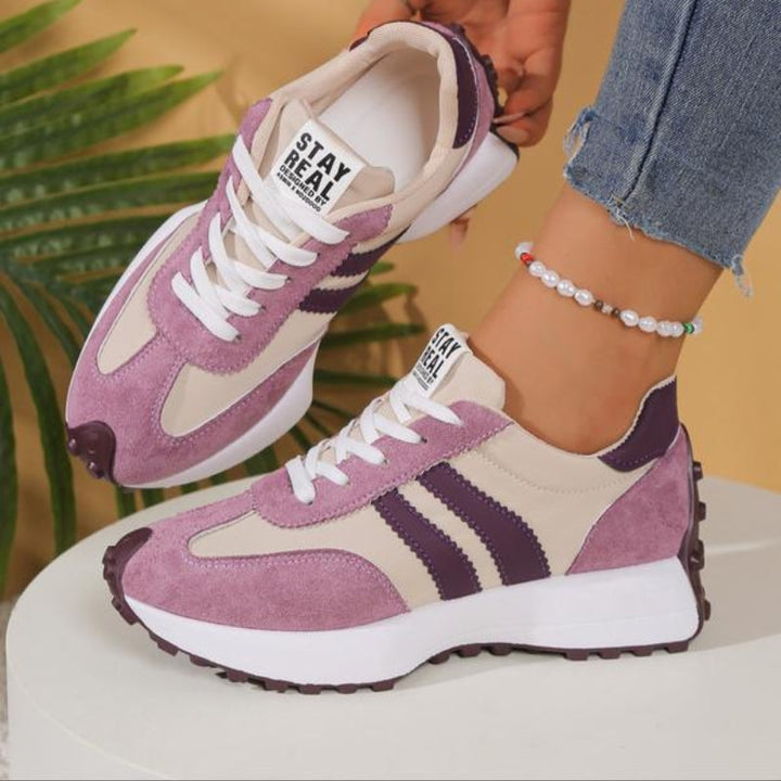 Różowo-beżowe sneakersy z fioletowymi paskami na stopach modelki stojącej na białym postumencie, obok liści palmy 11