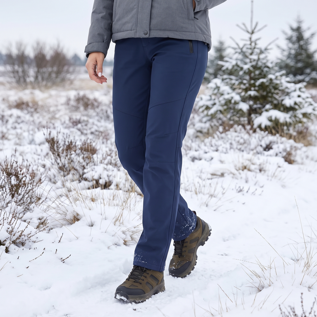 Granatowe spodnie na modelce idącej po ośnieżonym wrzosowisku, w szarej kurtce i brązowych butach trekkingowych 1