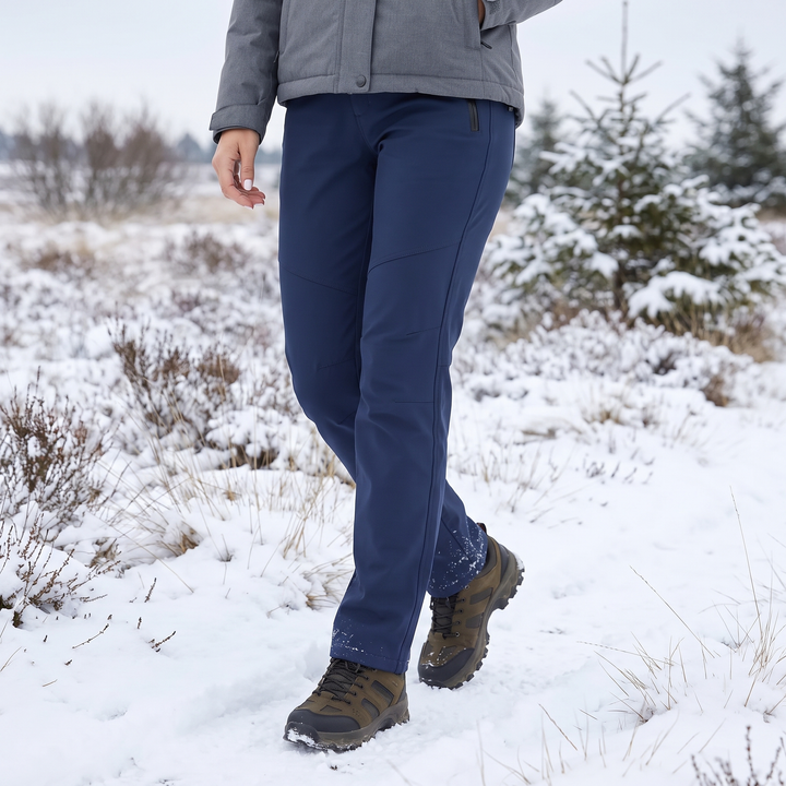 Granatowe spodnie na modelce idącej po ośnieżonym wrzosowisku, w szarej kurtce i brązowych butach trekkingowych 1