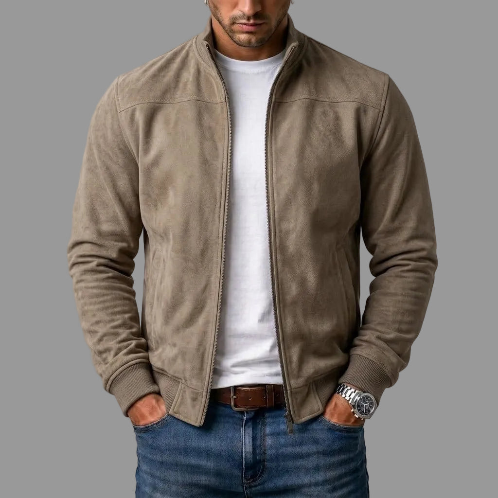 Mężczyzna w beżowo-szarej kurtce bomber ze stójką, rozpiętej na białym t-shircie, z rękami w kieszeniach jeansów. 2