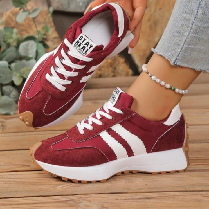 Bordowe sneakersy z białymi paskami na stopach modelki, na drewnianych deskach obok zielonych liści 4