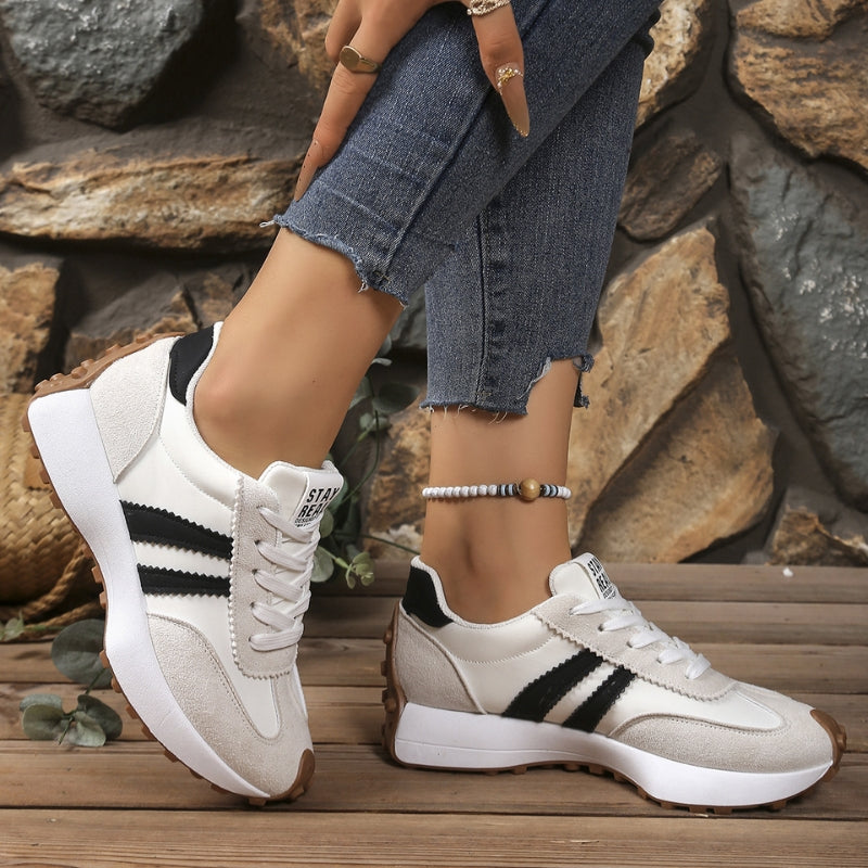 Białe sneakersy z beżowym zamszem i czarnymi paskami na nogach modelki, na tle kamiennej ściany 5
