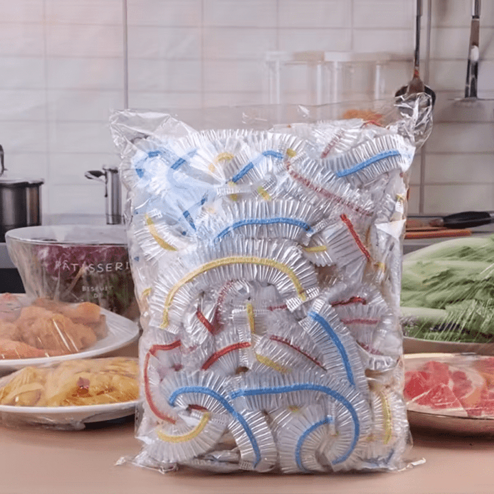 Przezroczysta torba pełna elastycznych pokrywek z kolorowymi paskami, ustawiona w kuchni obok talerzy z jedzeniem. 1