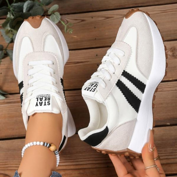 Białe sneakersy z czarnymi paskami i beżowym zamszem, jeden w dłoni, drugi na stopie, na drewnie 10