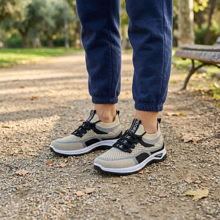 Stopy w beżowo-czarnych sneakersach z siateczki i granatowych spodniach dresowych na parkowej alejce z ławką w tle. 38
