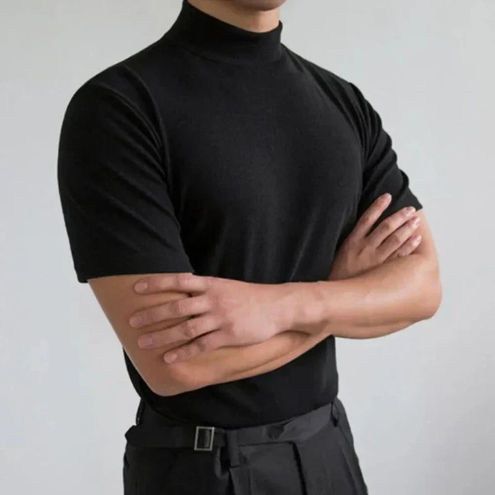 Ten sam czarny t-shirt z półgolfem na modelu ze skrzyżowanymi rękami, ujęcie na jasnoszarym tle. 2