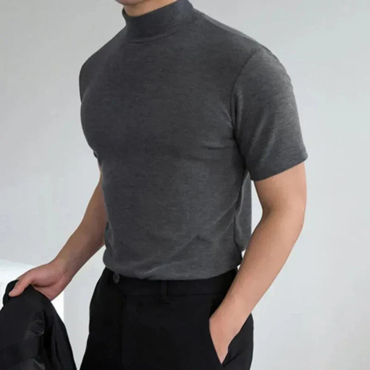Szary t-shirt z półgolfem na modelu z jedną ręką w kieszeni czarnych spodni, w drugiej trzyma ciemną marynarkę. 3