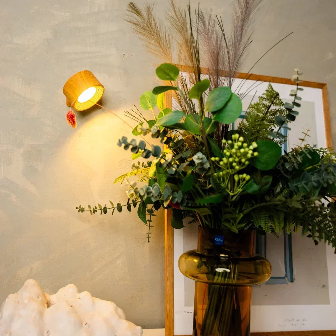 Lampa w drewnopodobnej obudowie oświetla ścianę nad bukietem sztucznych roślin w bursztynowym wazonie 5