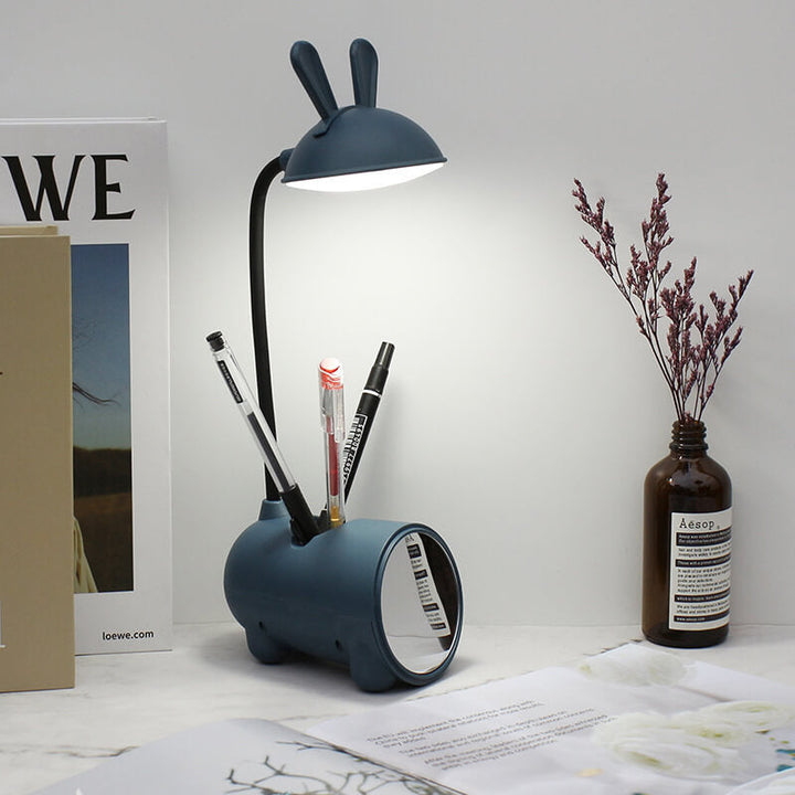 Granatowa lampka z uszkami świeci na biurku, w podstawie stoją długopisy, obok widać lusterko, szklaną butelkę z suszonymi kwiatami i książki. 9