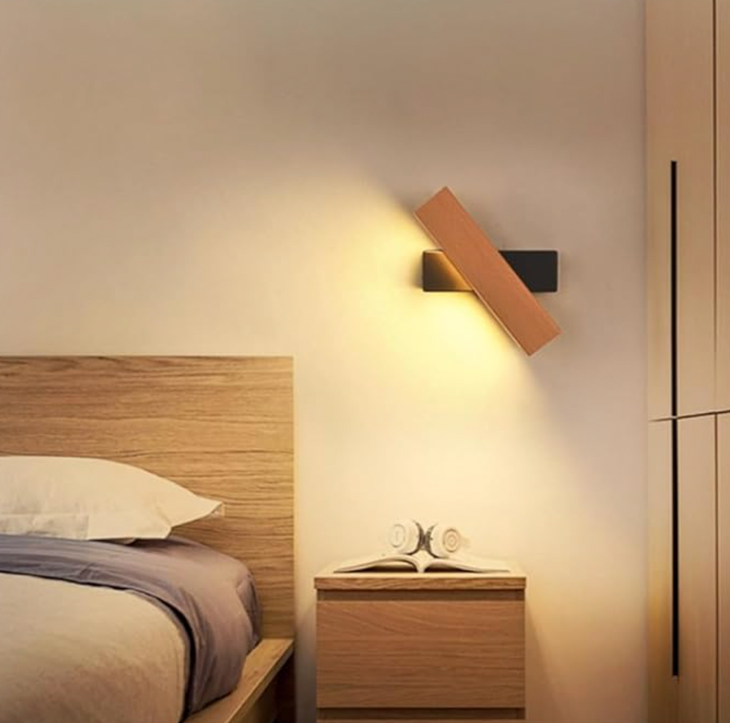 Wooden lamp on a black mount tilted diagonally above a bedside table with open book and headphones, next to a wooden bed headboard. 6