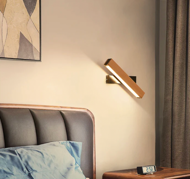 Wooden lamp on a black mount above a bedside table with digital clock, beside a grey padded headboard and curtain. 8