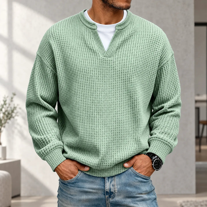 Miętowozielony sweter waflowy z wycięciem przy dekolcie na modelu w jeansach, na tle jasnego wnętrza, ręce w kieszeniach. 1