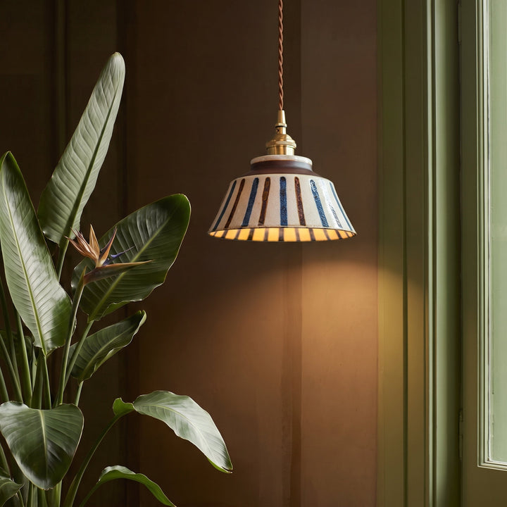 Lampa w pasy z mosiężną oprawą obok dużej rośliny o zielonych liściach, w ciepło oświetlonym wnętrzu z brązową ścianą. 4