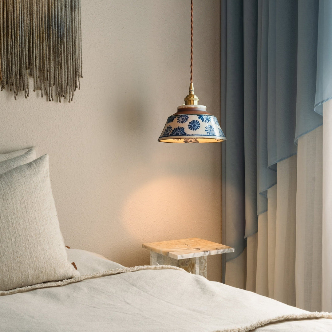 Lampa w niebieskie kwiaty świeci nad stolikiem z jasnego kamienia obok łóżka z lnianą pościelą i błękitną zasłoną. 2