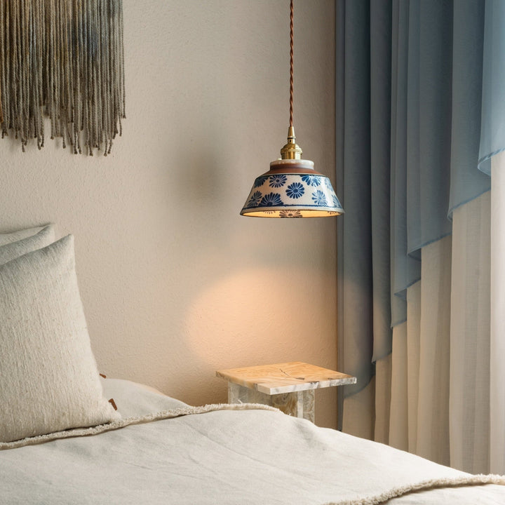 Lampa w niebieskie kwiaty świeci nad stolikiem z jasnego kamienia obok łóżka z lnianą pościelą i błękitną zasłoną. 2
