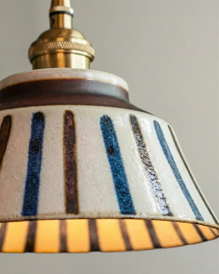 Zbliżenie na ceramiczny abażur w niebiesko-brązowe pasy ze szkliwem i mosiężną oprawką, wnętrze świeci ciepłym światłem. 7