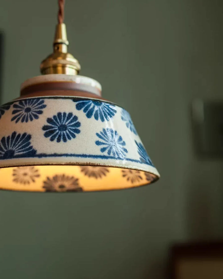 Zbliżenie na ceramiczny abażur z niebieskimi kwiatami, mosiężną oprawką i skręconym przewodem, widać szkliwioną fakturę. 6
