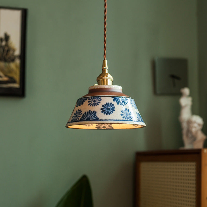Lampa z abażurem w niebieskie kwiaty, zawieszona na skręconym brązowym przewodzie, na tle zielonej ściany z obrazem i rzeźbą. 1