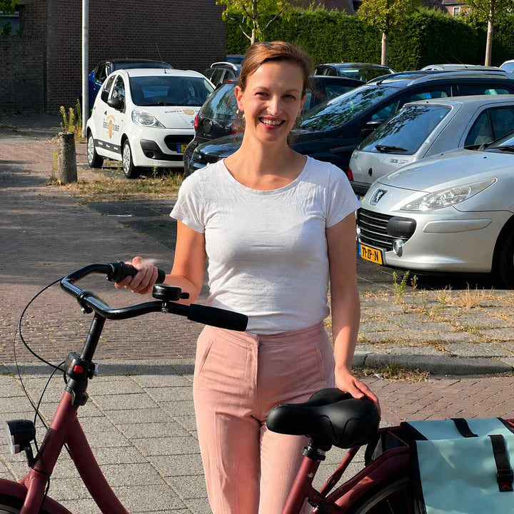 Uśmiechnięta kobieta stoi obok roweru miejskiego z zamontowanym czarnym siodełkiem i niebieskimi sakwami. 7