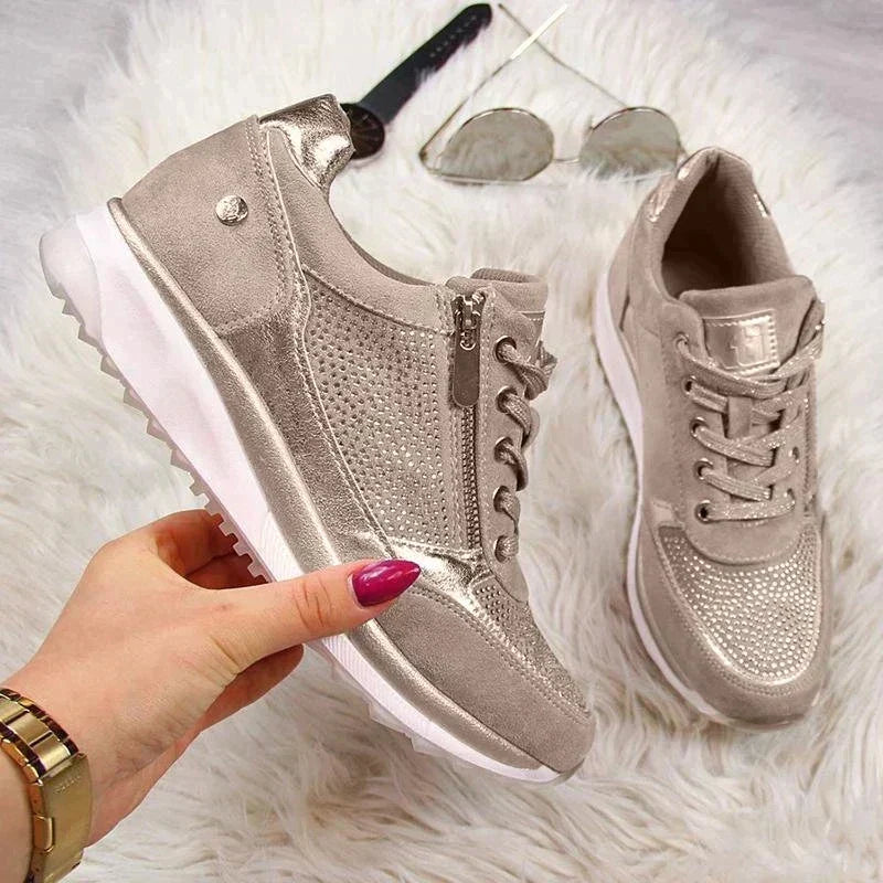 Dłoń z różowymi paznokciami trzyma beżowy sneaker z zamkiem i srebrzystym połyskiem, drugi but obok na futrzanym tle z okularami. 7