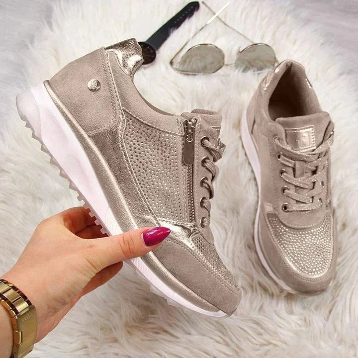 Dłoń z różowymi paznokciami trzyma beżowy sneaker z zamkiem i srebrzystym połyskiem, drugi but obok na futrzanym tle z okularami. 7