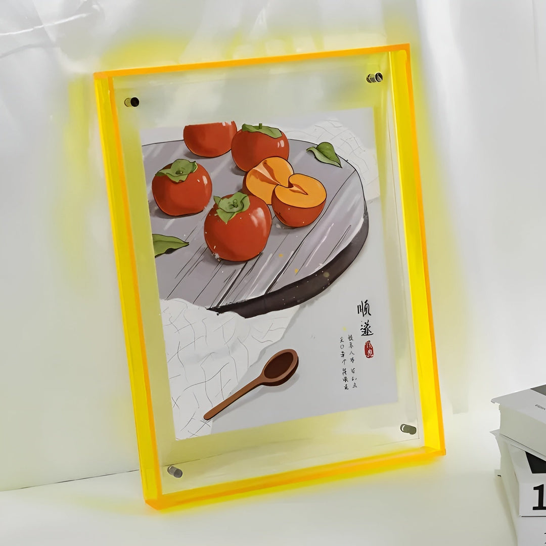 Żółta akrylowa ramka z ilustracją persymonów na talerzu i drewnianą łyżką, oparta o ścianę obok książek. 12