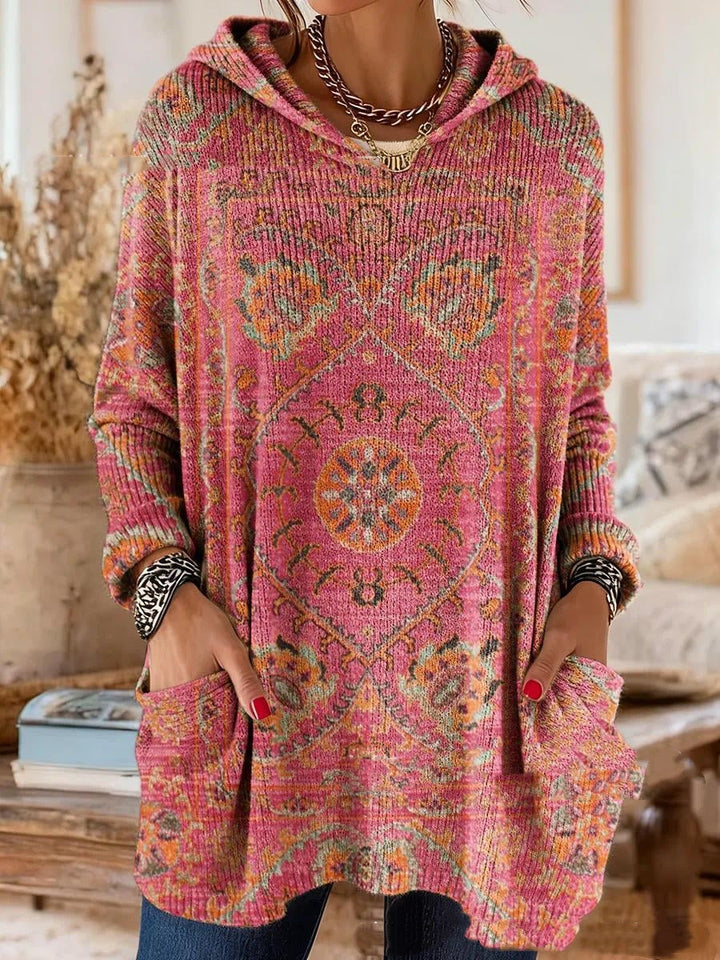 Różowa tunika z kapturem z pomarańczowo-zielonym ornamentem w stylu boho, dzianina w prążki, modelka trzyma dłonie w kieszeniach. 6