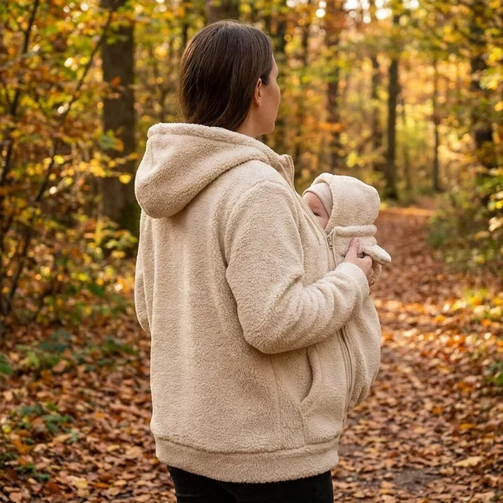 Widok z boku: kobieta w beżowej puchatej bluzie stoi w jesiennym lesie i niesie niemowlę w panelu z kapturkiem. 6