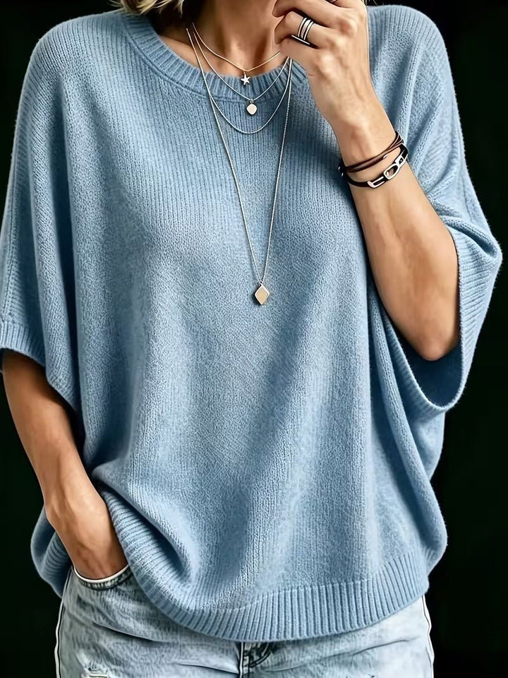 Niebieski dzianinowy top z rękawami batwing, zbliżenie z przodu na modelce z naszyjnikami, ręka w kieszeni dżinsów. 12