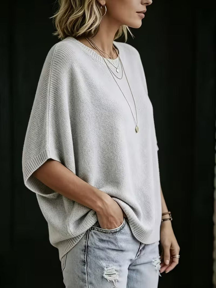 Jasnoszary top z dzianiny widziany z boku, luźny krój i szerokie rękawy batwing, modelka z ręką w kieszeni dżinsów. 2