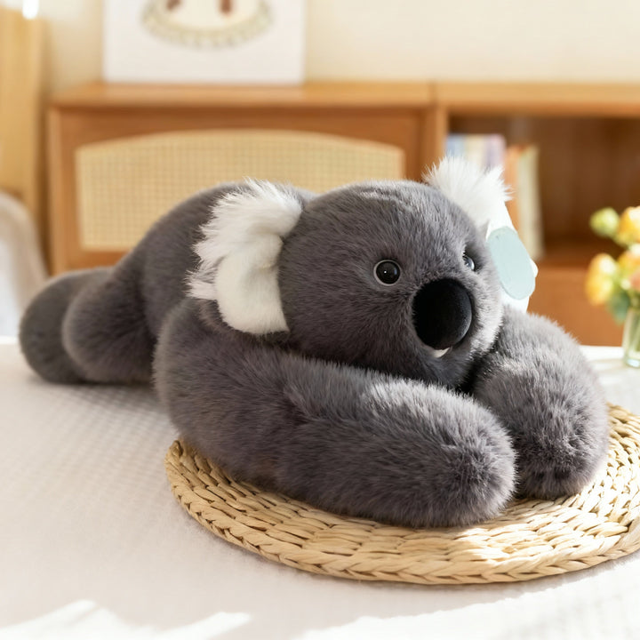 Szara pluszowa koala z białymi uszami i dużym czarnym nosem leży na słomianej podkładce, a długie ramiona obejmują jej głowę. 16
