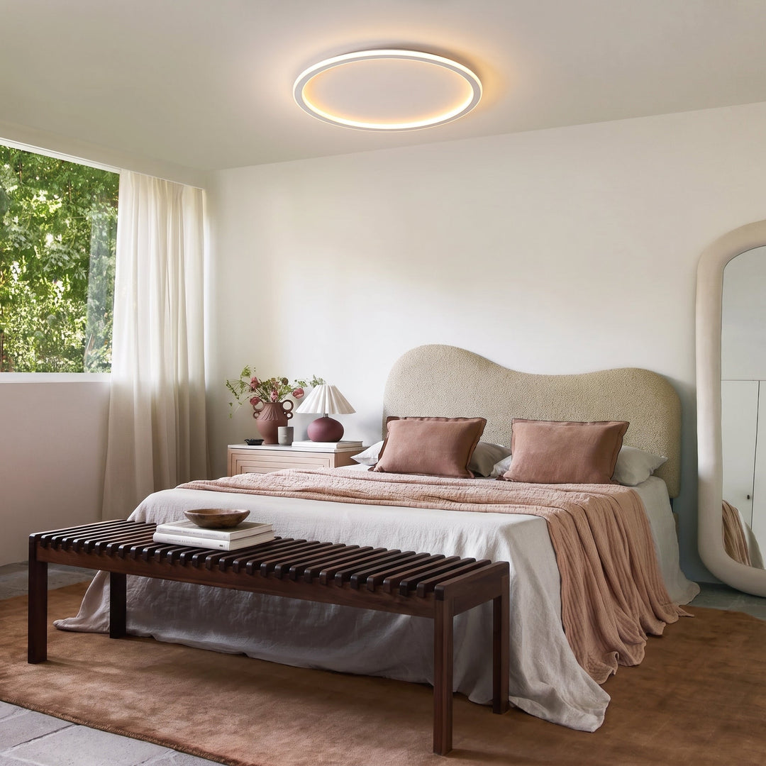 Lampa pierścieniowa nad łóżkiem w eleganckiej sypialni z różowymi poduszkami, drewnianą ławką i dużym lustrem. 11